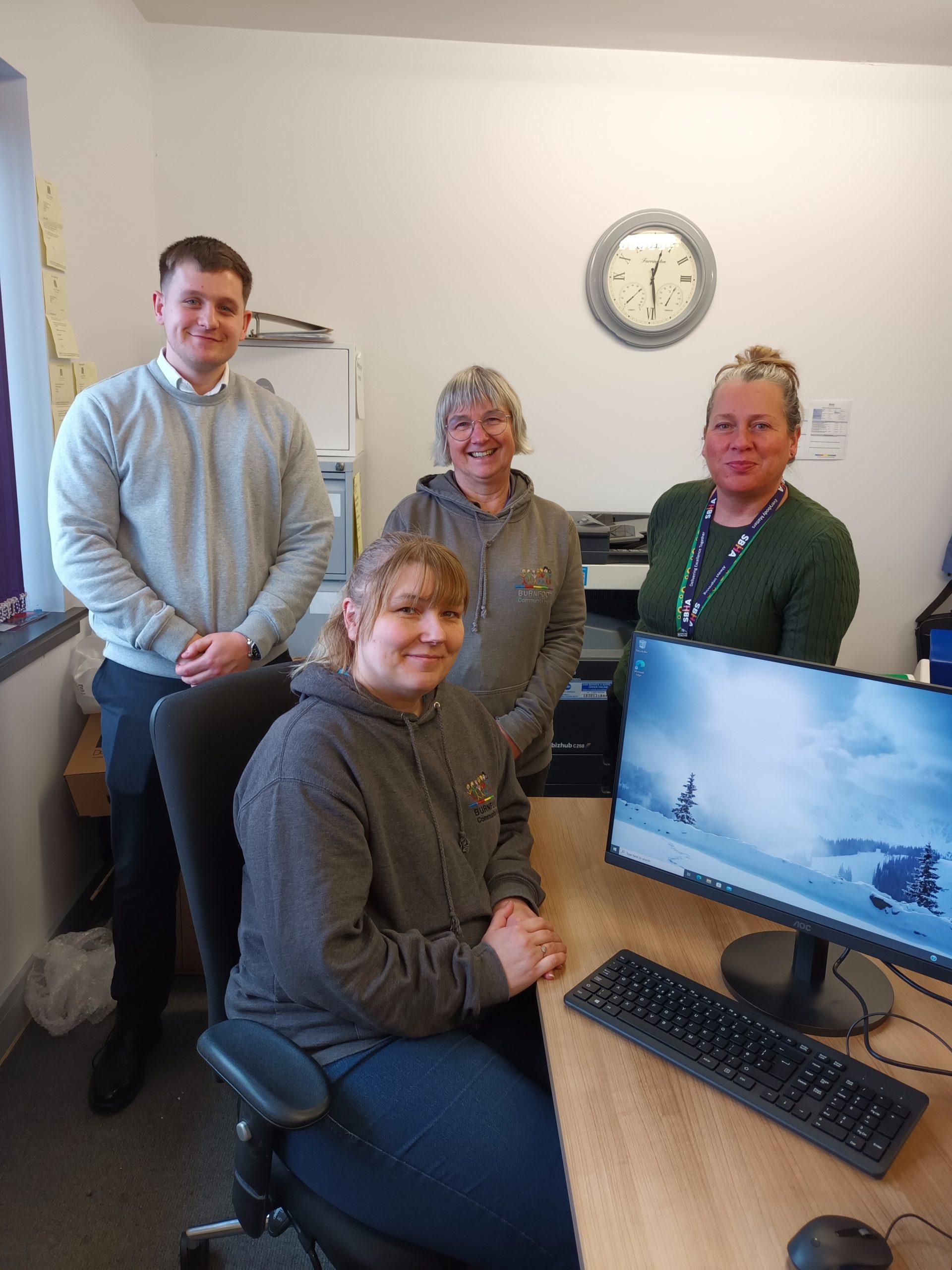 A group of four people posing inside an office. One person is seated at a wooden desk with a computer monitor displaying a snowy landscape, while the other three stand behind them. Two of the standing individuals are wearing casual hoodies with a colorful logo, while another is wearing a green sweater with a lanyard, and the fourth person is dressed in a light gray sweater. The office has a clock on the wall, filing cabinets, and various papers attached to the walls. The atmosphere is friendly and welcoming.