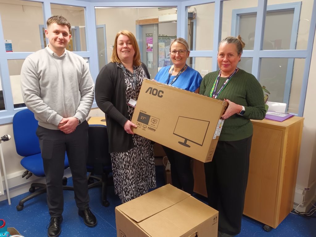 A group of four people standing in an office setting with blue flooring and windows dividing the space. Two women in the center are holding large cardboard boxes labeled "AOC" containing 27-inch monitors. One woman is wearing a blue uniform, another is wearing a patterned dress and black cardigan, and a third is wearing a green sweater with a lanyard. A man in a light gray sweater stands on the left, smiling. Additional boxes are stacked on the floor, and blue office chairs and a wooden cabinet are visible in the background. The atmosphere is positive and professional.